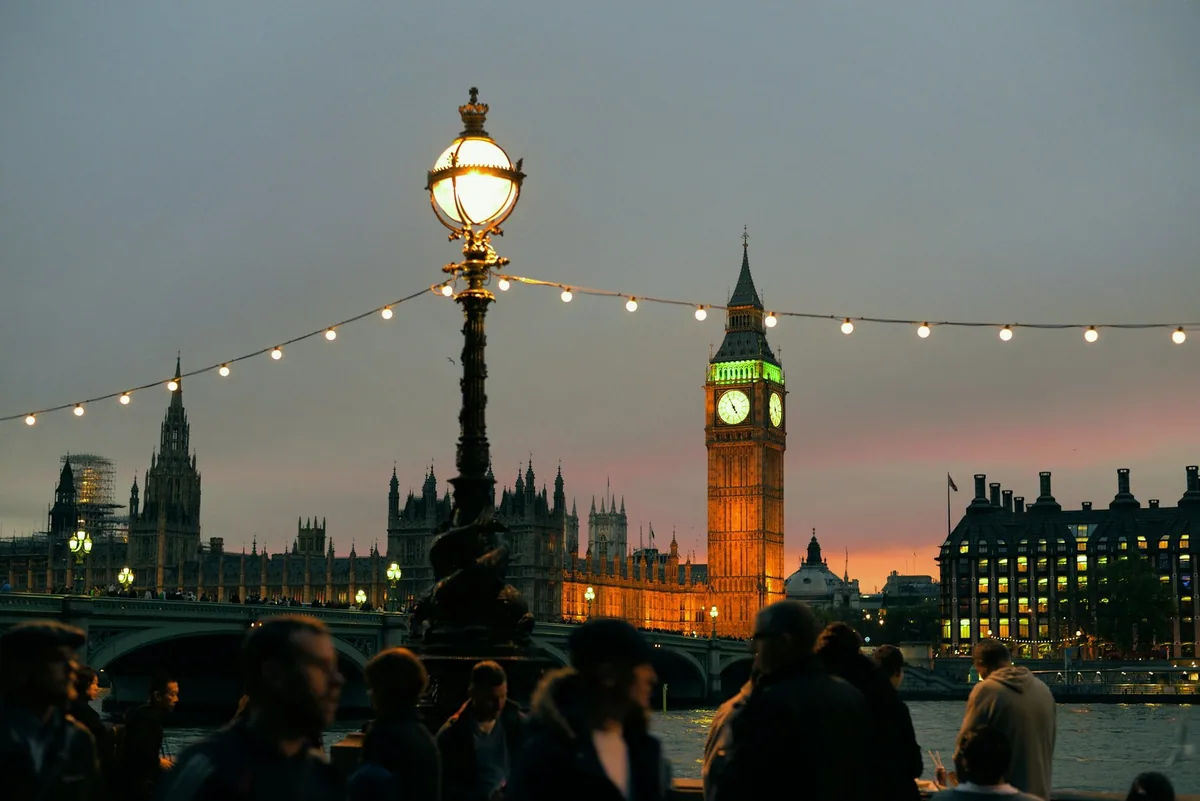 Folk på elvepromenaden i skumringen, med lyktestolpe, lysslynger og et opplyst Big Ben på andre siden av Themsen