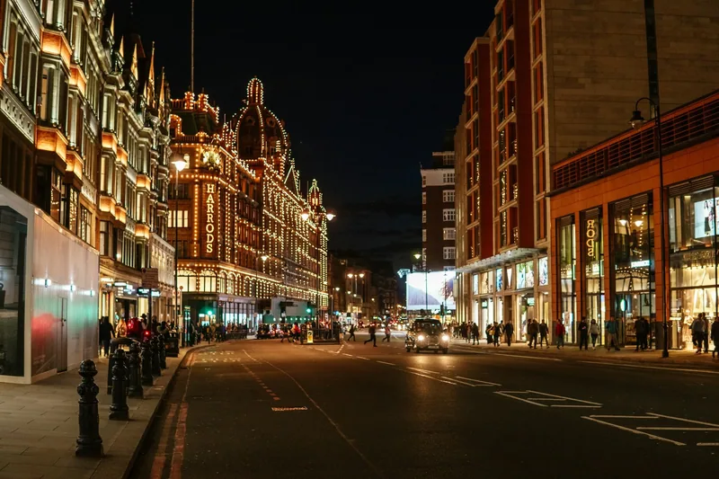Den opplyste fasaden til Harrods i Brompton Road om kvelden, med folk på fortauene og en taxi i gata