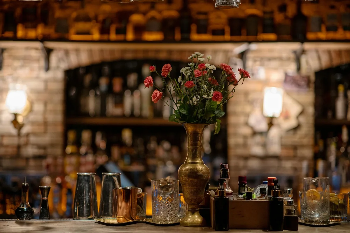 Dunkel cocktailbar med murvegg, vegglamper og en messingvase med roser foran flaskehyllene