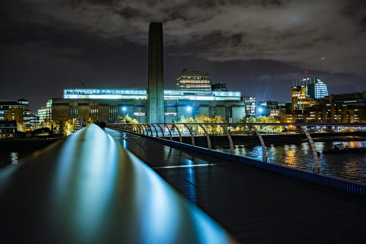 Tate Modern med det høye skorsteinstårnet, opplyst om kvelden og sett fra Millennium Bridge over Themsen