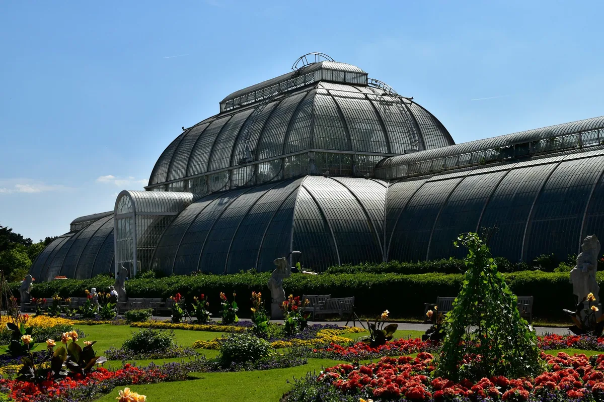 Palm House i Kew Gardens, et viktoriansk veksthus i glass og jern, med blomsterbed foran en solfylt dag