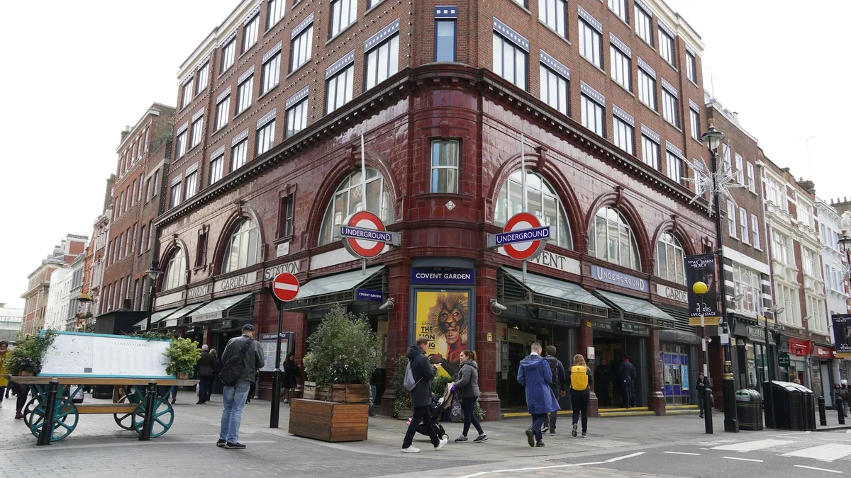 Den rødbrune fasaden til Covent Garden stasjon med undergrunnsskilt, og folk som går forbi på gaten utenfor