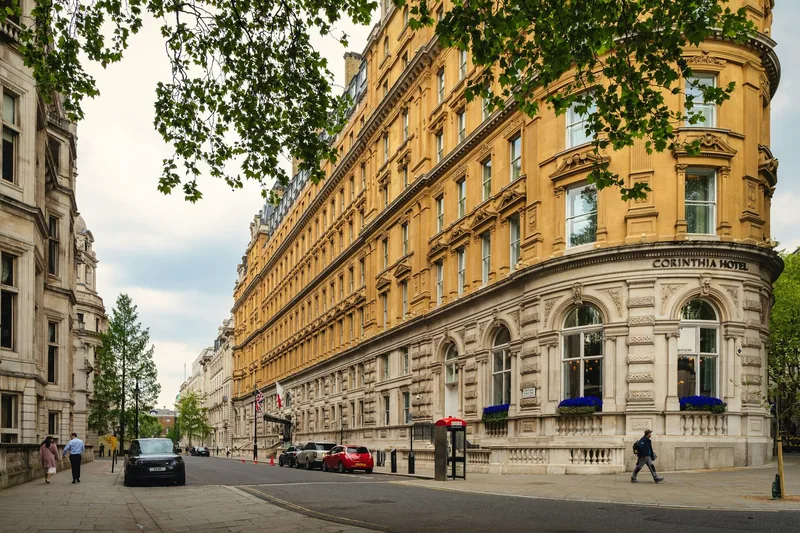 Den buede gule fasaden til Corinthia Hotel i en rolig gate i Westminster under grønne trekroner