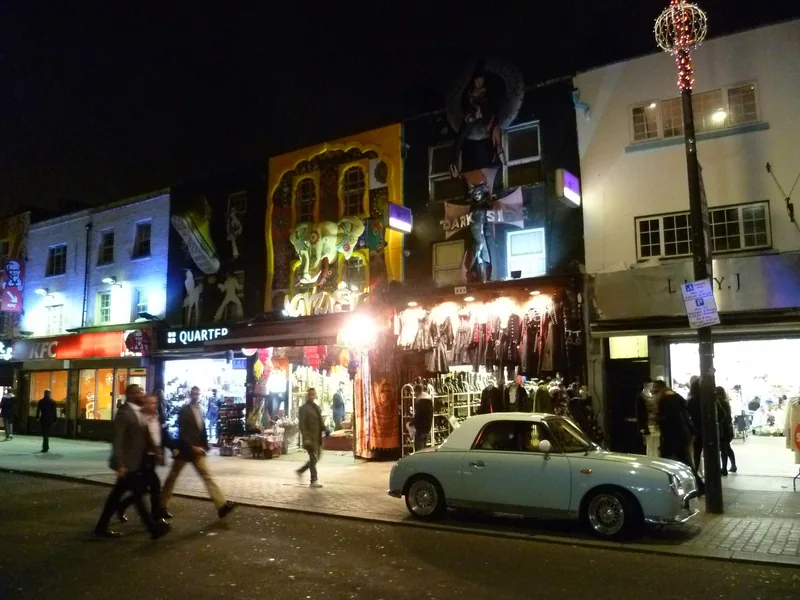Butikker med store fasadefigurer og opplyste vinduer i Camden Town en kveld, med fotgjengere og en lyseblå bil foran