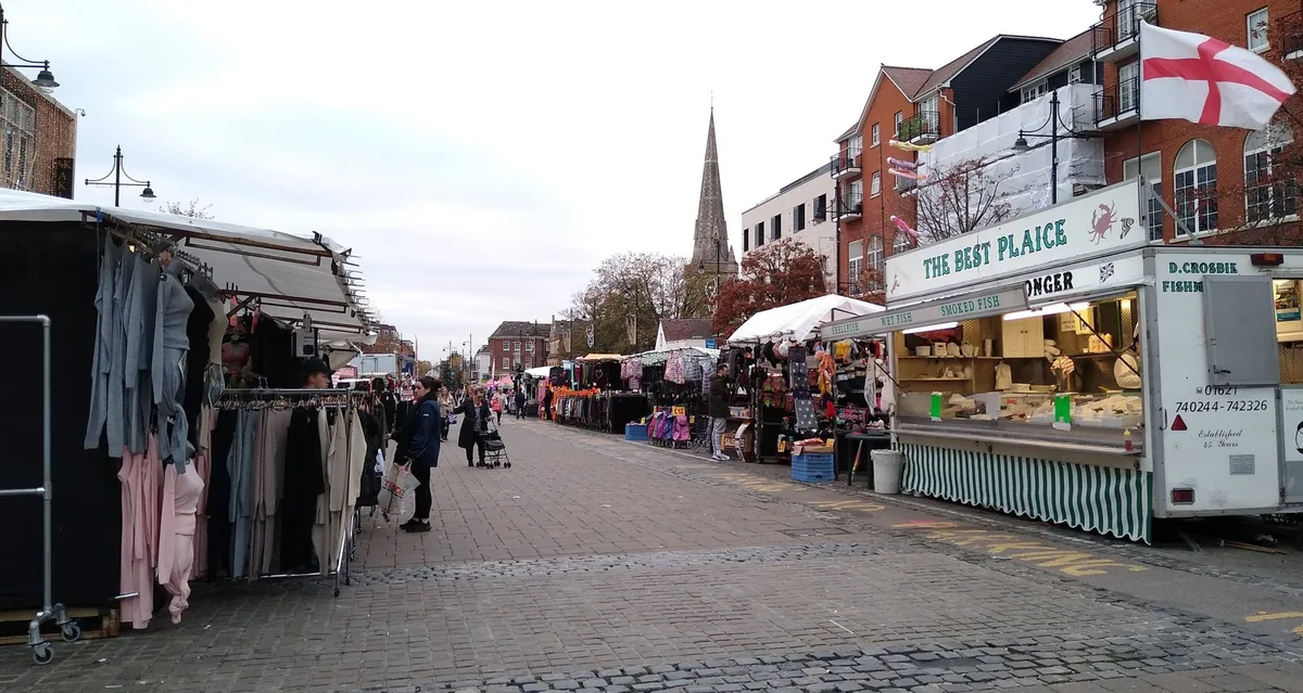 Romford Market med klesboder og en fiskevogn langs en brolagt plass, og et kirkespir i bakgrunnen