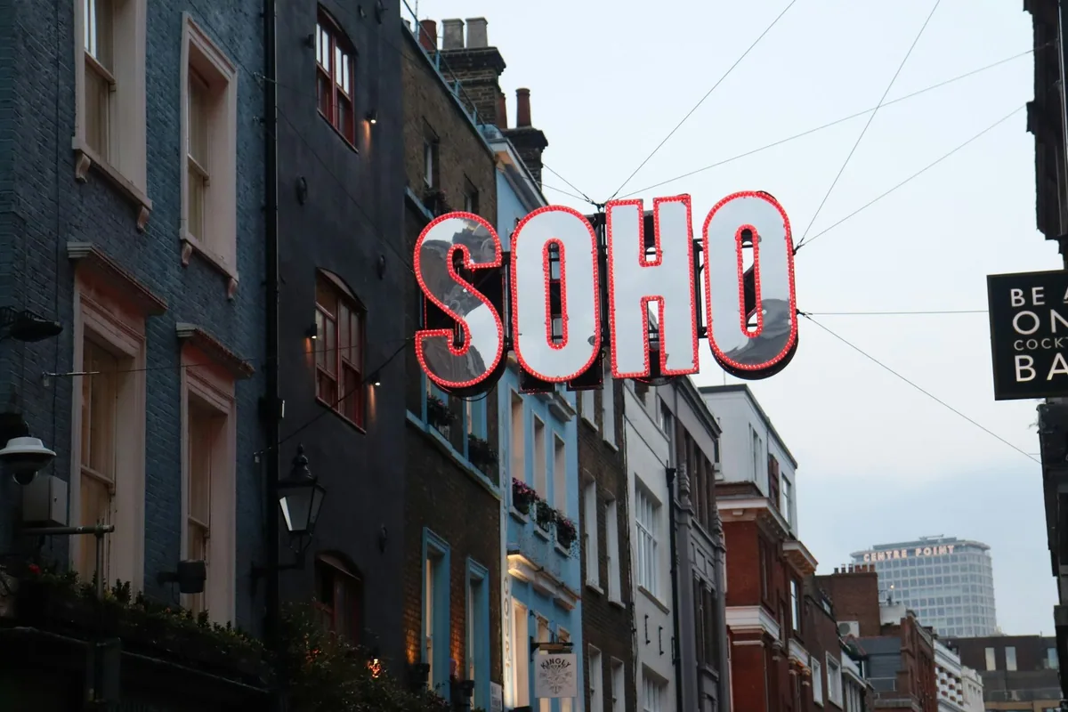 Store SOHO-bokstaver med røde lyspærer henger over en smal gate i Soho, med Centre Point i bakgrunnen