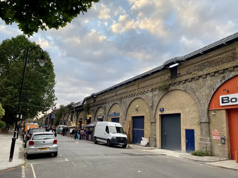 Jernbanebuer i gul murstein i Bermondsey, med små barer og folk samlet langs gata en sommerkveld