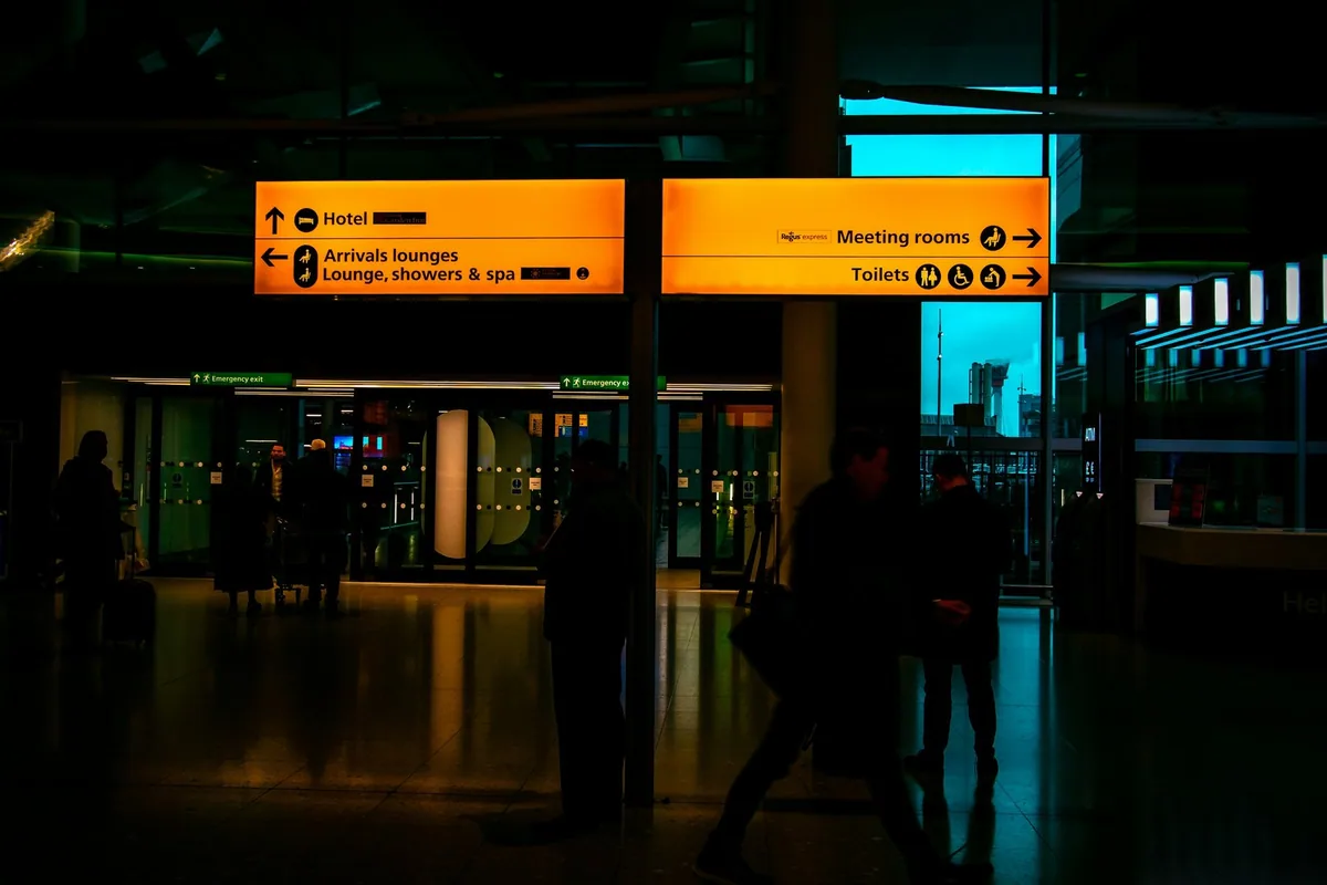 Gule skilt til ankomstlounger, hotell og toaletter i en flyplassterminal, med silhuetter av reisende