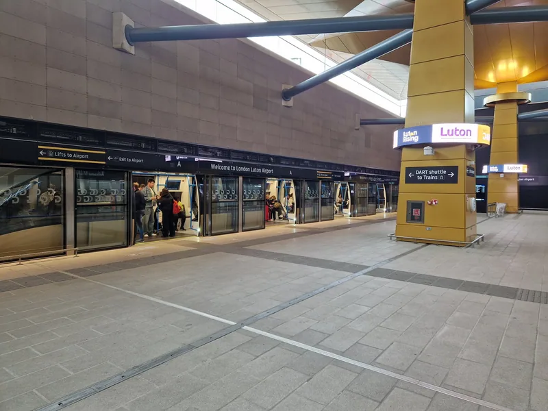 Perrongen på DART-stasjonen under London Luton Airport, med skyttelbanen og skilt mot heisene til terminalen