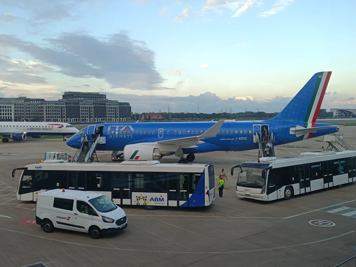 Passasjerer går om bord i et blått fly på London City Airport, med flyplassbusser foran og bygninger bak