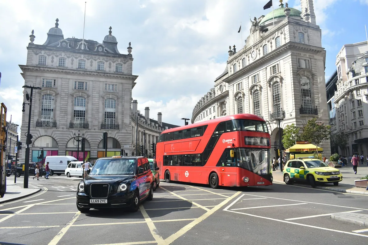 Rød dobbeltdekkerbuss og svart taxi i Piccadilly Circus, omgitt av klassiske fasader