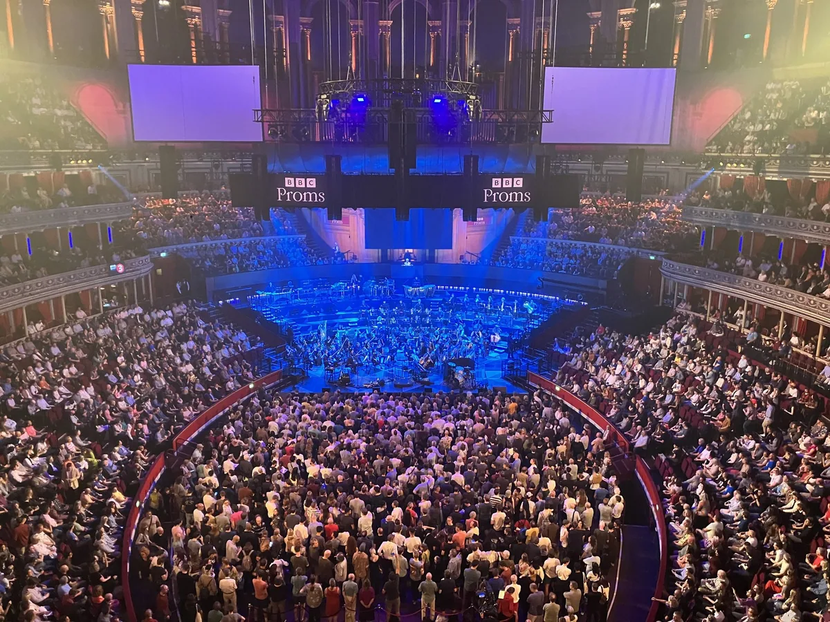 Proms-konsert i Royal Albert Hall sett fra balkongen, med orkesteret i blått lys og ståplasspublikum i arenaen