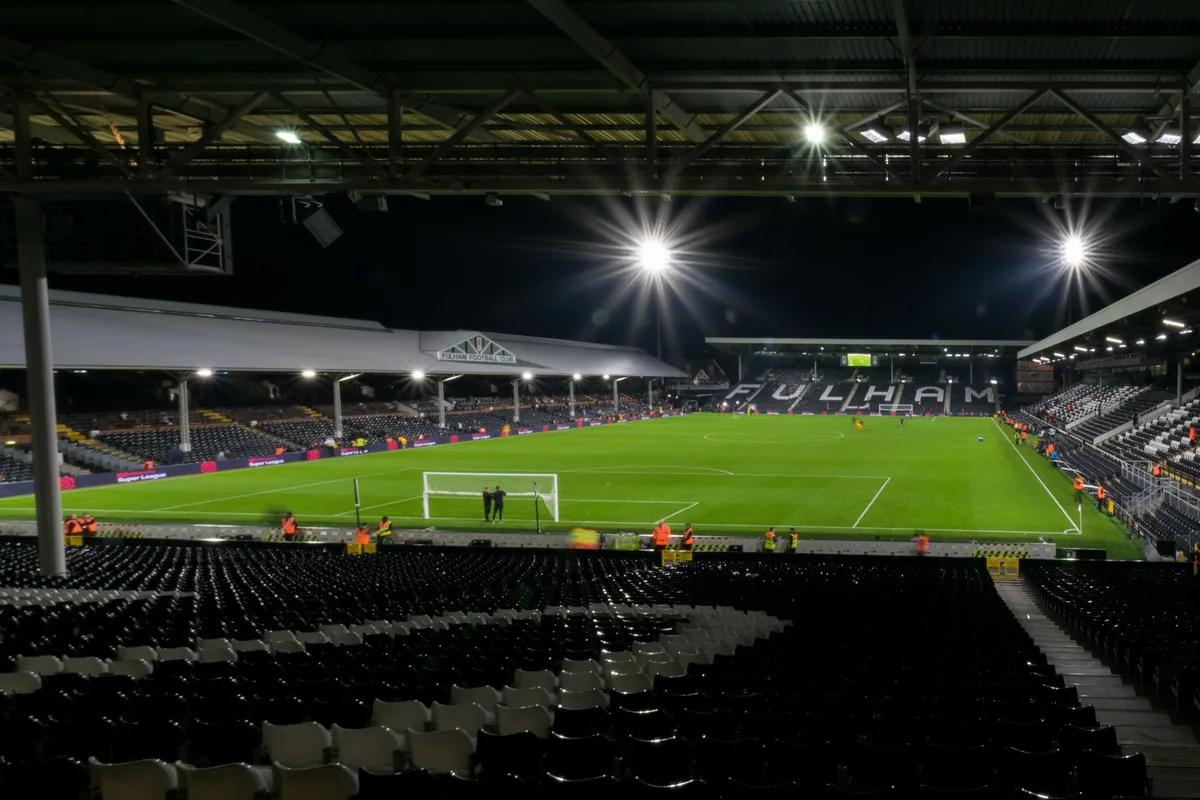 Craven Cottage i flomlys en kveld, med tomme seter i forgrunnen og FULHAM skrevet på tribunen bak målet
