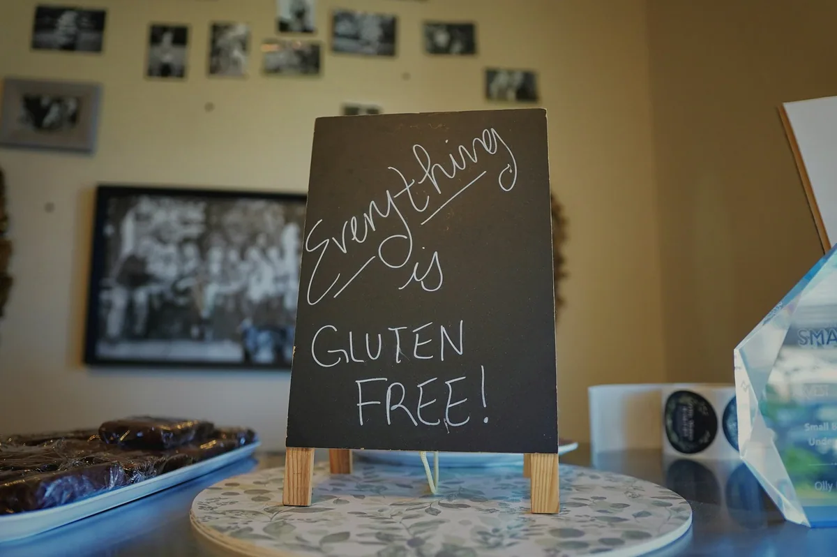 Liten tavle på en kafédisk med håndskrevet tekst om at alt er glutenfritt, ved siden av et fat med brownies