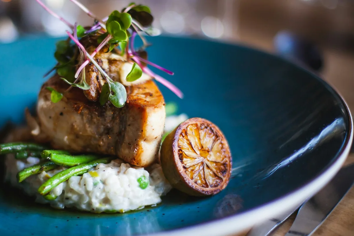 Stekt fiskefilet med mikrogrønt, aspargesbønner, risotto og grillet sitron i en blå tallerken