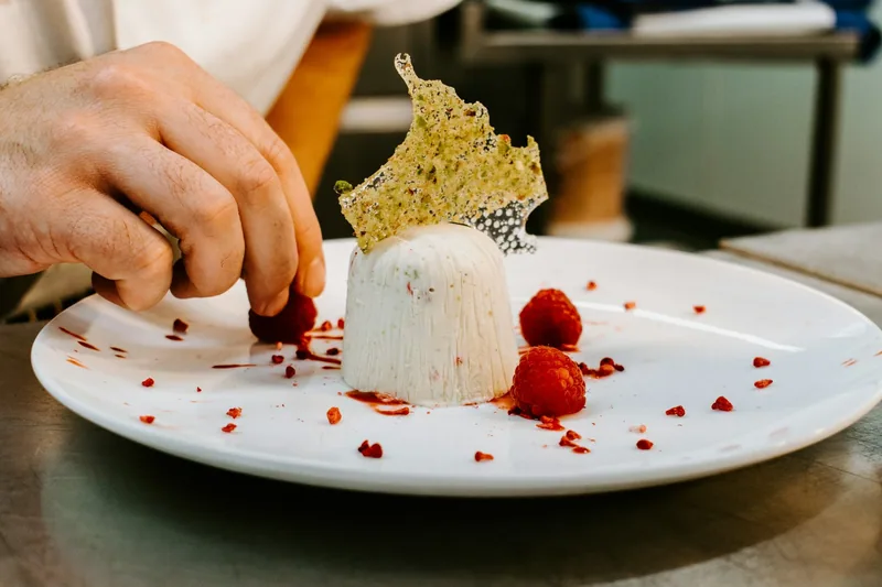 Kokk legger bringebær rundt en hvit dessert med en tynn nøttekjeks på toppen, på en benk i restaurantkjøkkenet