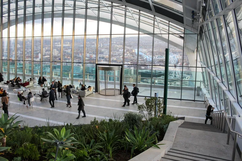 Gjester ved bord og sofaer inne i glasskuppelen i Sky Garden, med utsikt over London gjennom vinduene