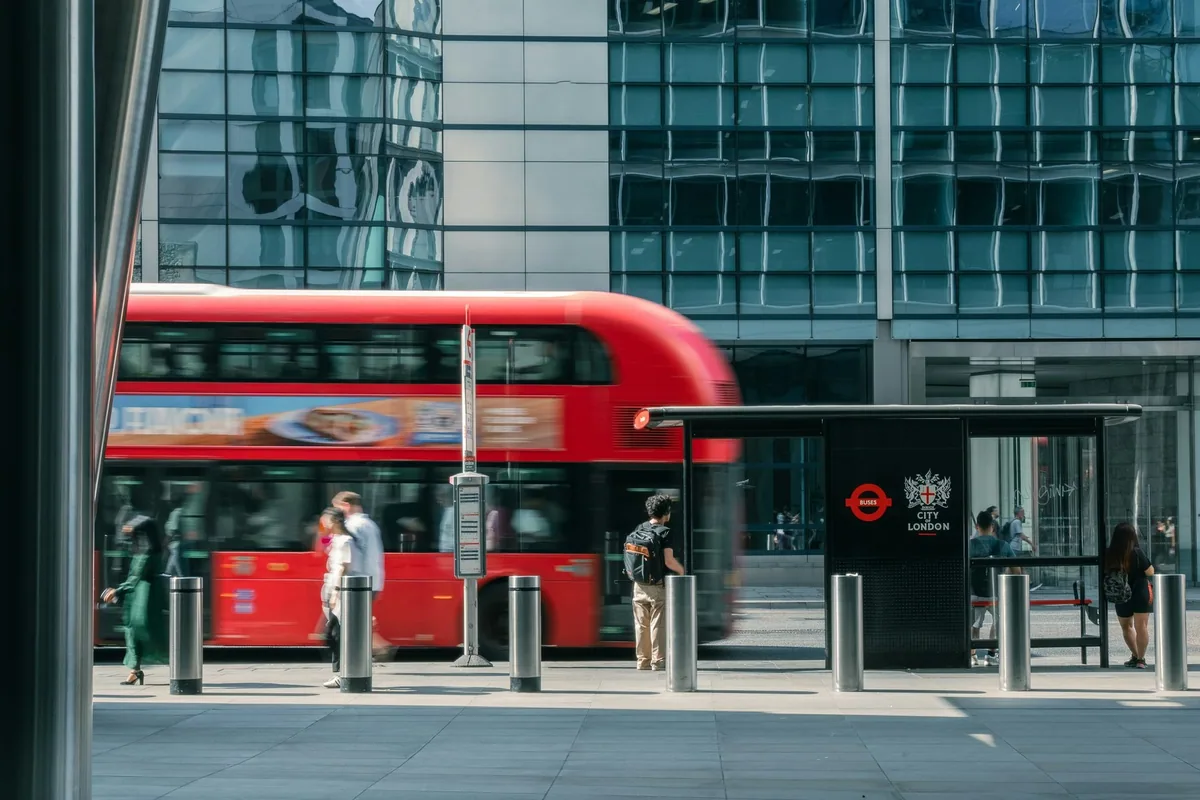 Rød dobbeltdekker passerer et busskur i City of London der noen reisende venter