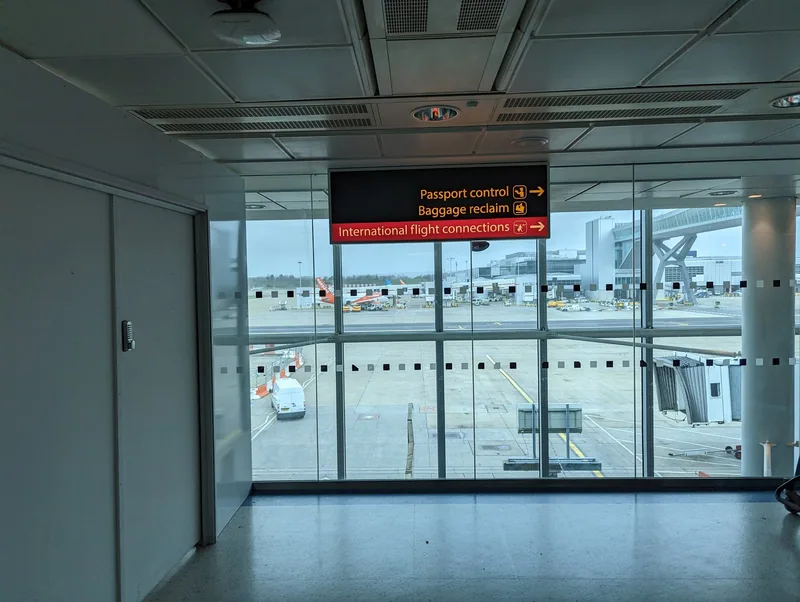 Skilt mot passkontroll og bagasjebånd i en ankomstkorridor på Gatwick, med flyoppstillingsplassen bak glasset