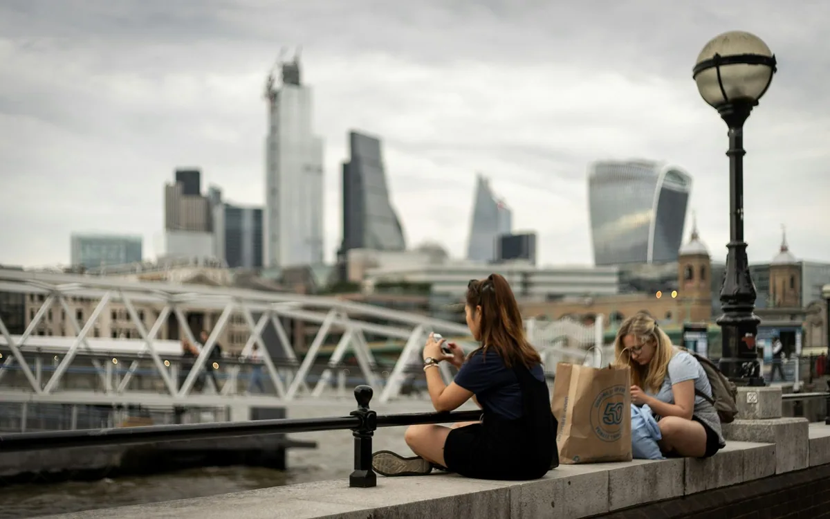 To kvinner med handlepose sitter på muren ved Themsen, med skyskraperne i City of London i bakgrunnen