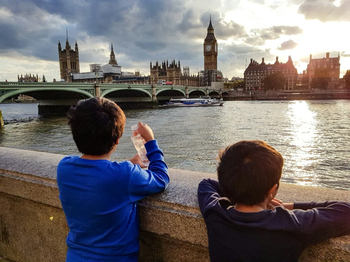 To gutter ved muren langs Themsen ser ut over elven mot Westminster Bridge og en elvebåt i kveldssol