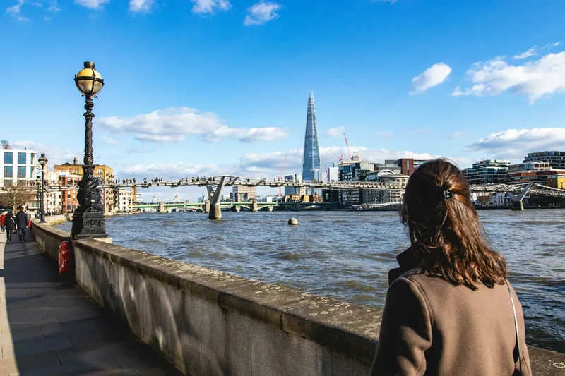 Kvinne sett bakfra på elvepromenaden, med Millennium Bridge og The Shard på andre siden av Themsen