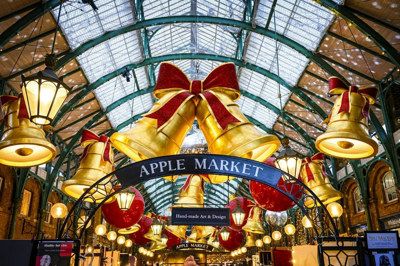 Store gullklokker med røde sløyfer henger under glasstaket i Apple Market i Covent Garden i juletiden