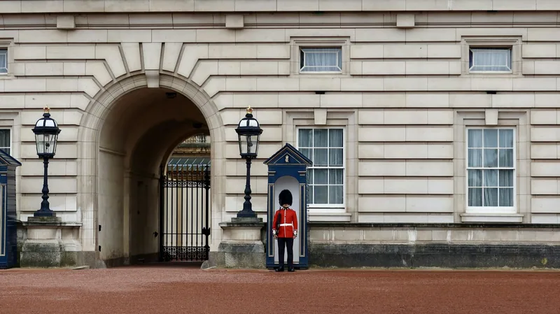 Vaktsoldat i rød uniform og bjørneskinnslue foran vaktbua ved Buckingham Palace
