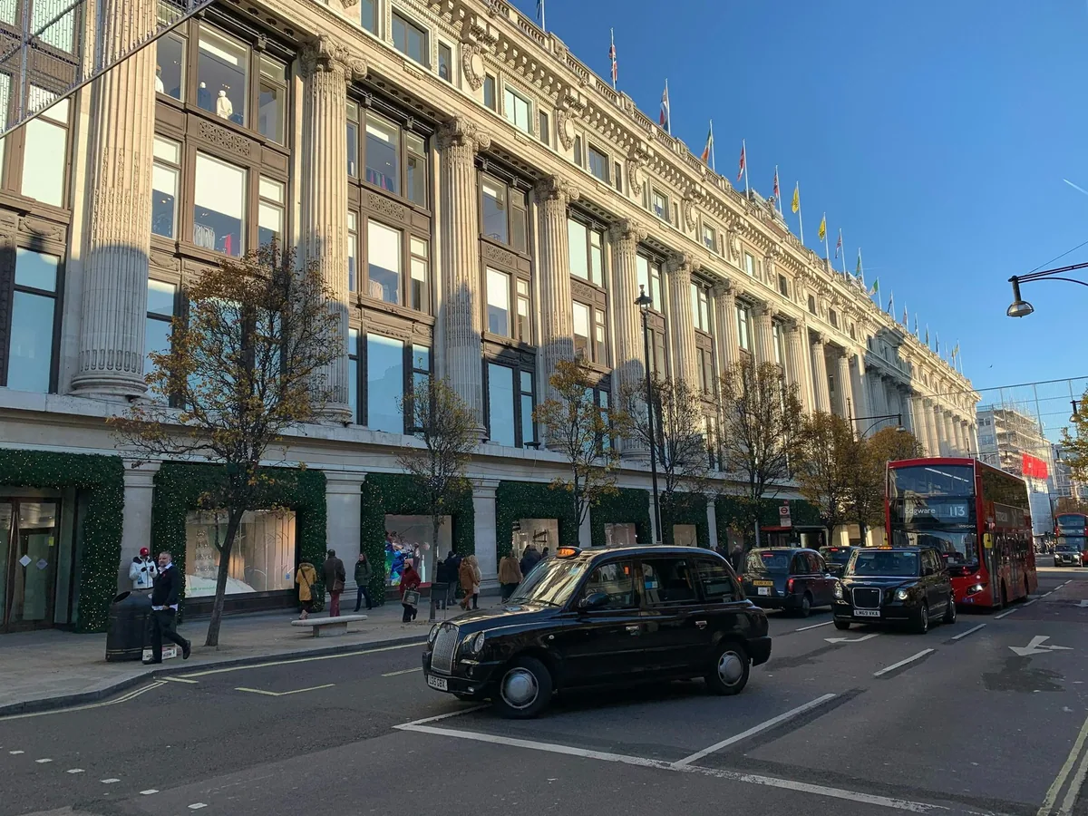 Søylefasaden til varehuset Selfridges i Oxford Street, med svarte taxier og en rød buss i gata