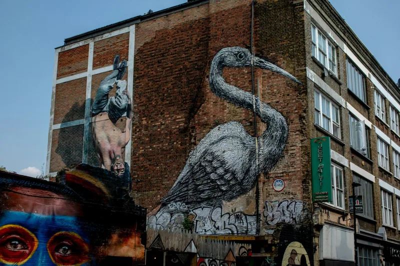 Stort veggmaleri av en grå fugl på en murvegg i Brick Lane-strøket, med annen gatekunst rundt