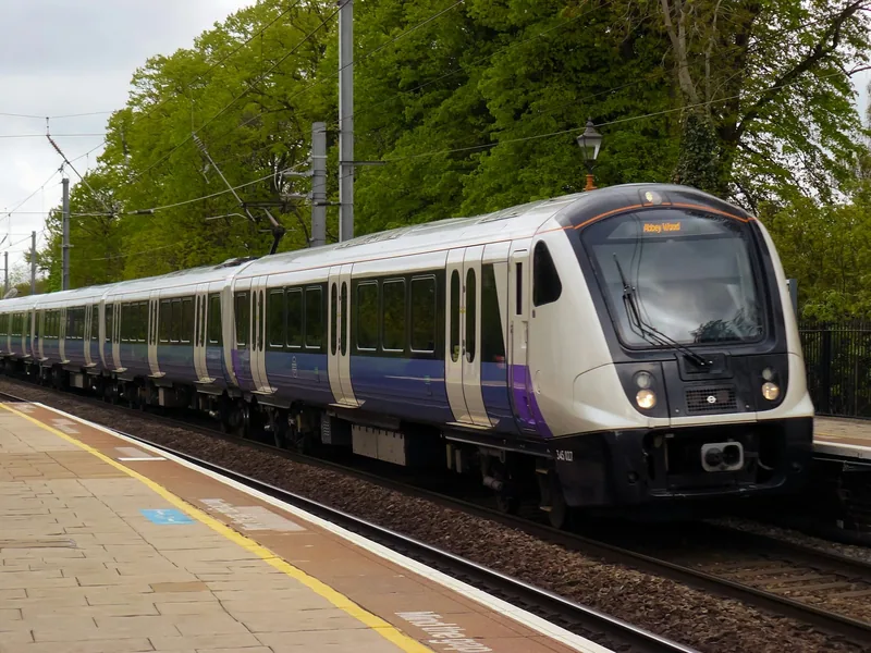Lilla og hvitt Elizabeth line-tog kjører inn på en perrong omgitt av grønne trær