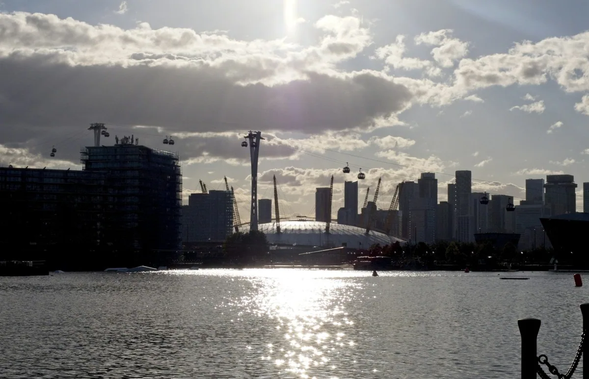 Kabelbanens gondoler og master i motlys over vannet, med The O2 og skyskraperne i Canary Wharf bak