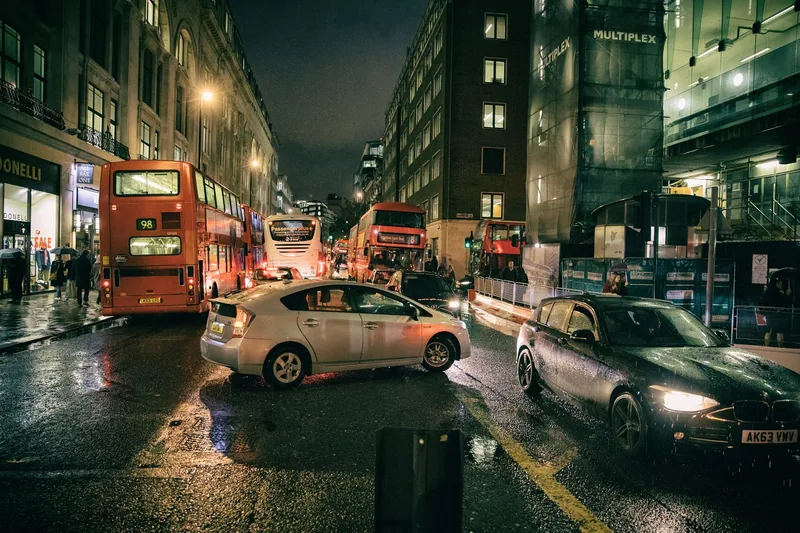 Biler og røde dobbeltdekkere i kveldstrafikken på en regnvåt New Oxford Street